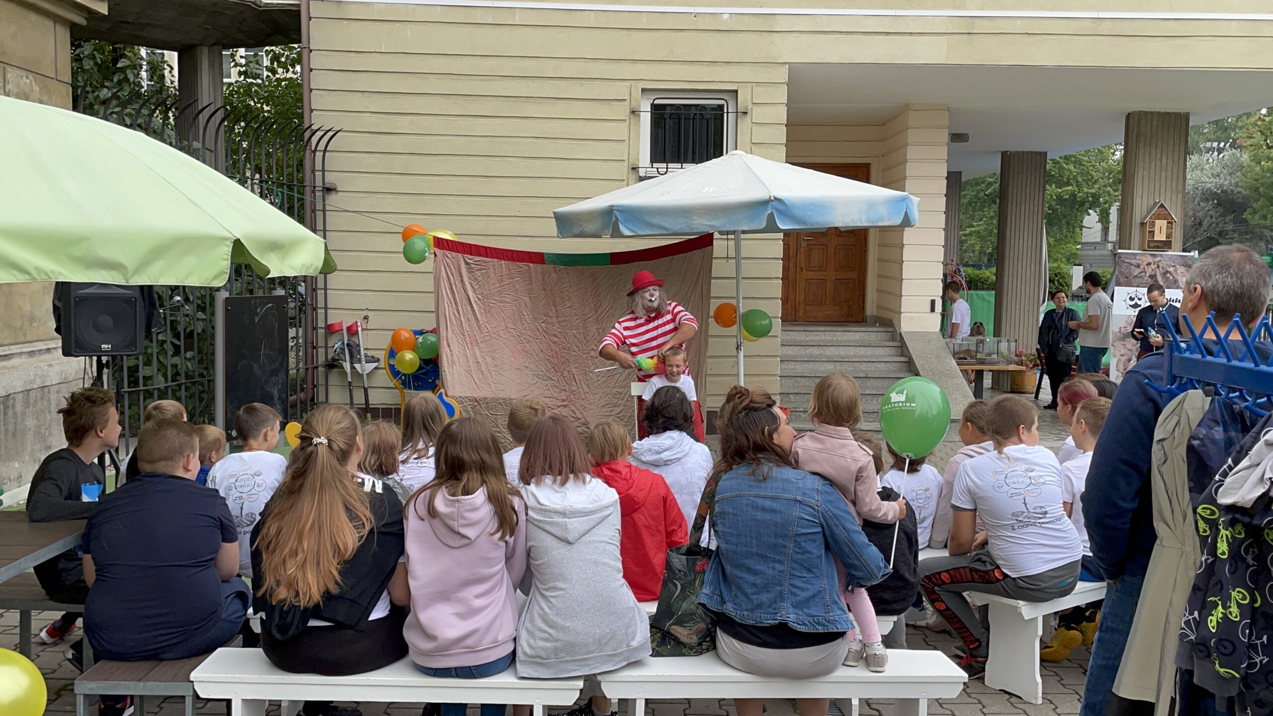 Występ klauna. Klaun w czerwono-białym stroju bawi widownię złożoną z dzieci i dorosłych siedzących na ławkach.