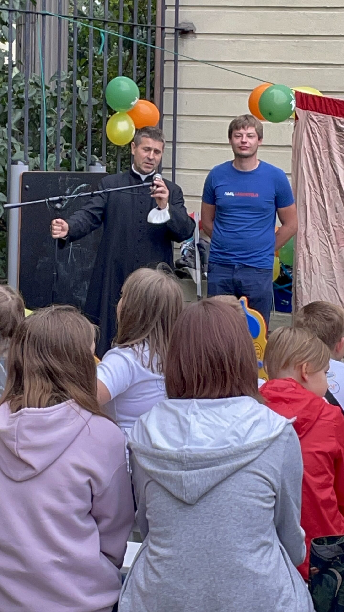 Słowo do dzieci. Prowadzący przemawia do mikrofonu, przed nim siedzi grupa dzieci, w tle balony.