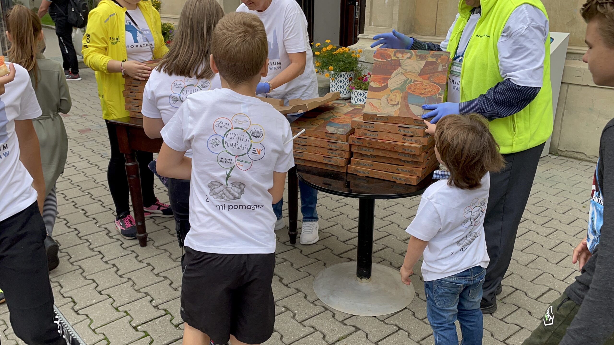 Wspólny poczęstunek. Wydawanie pizzy na placu, wokół stołów zebrały się dzieci i dorośli.