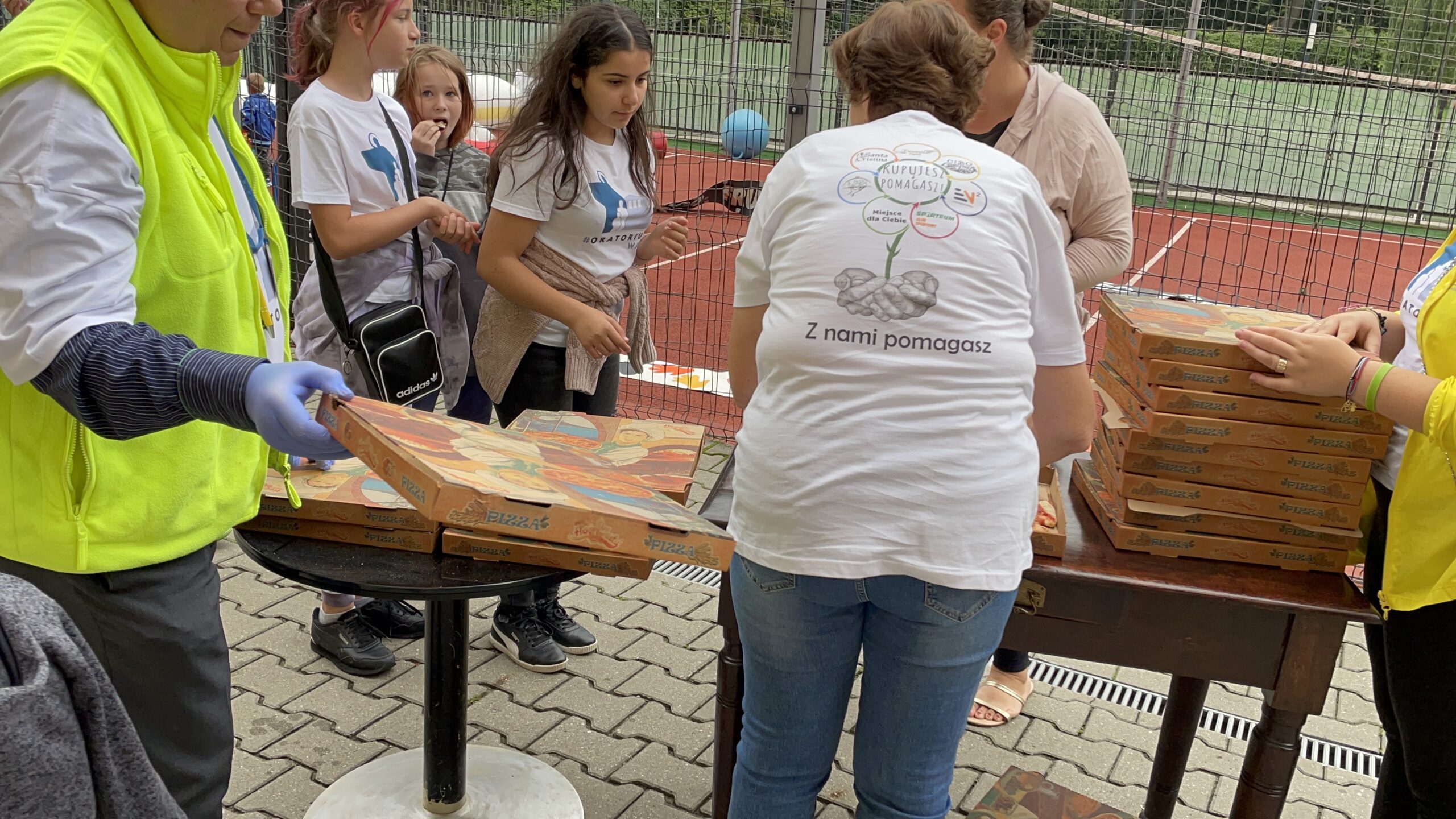Pizza dla wszystkich. Stoliki z kartonami pizzy przy boisku, dorośli i dzieci czekają na poczęstunek.