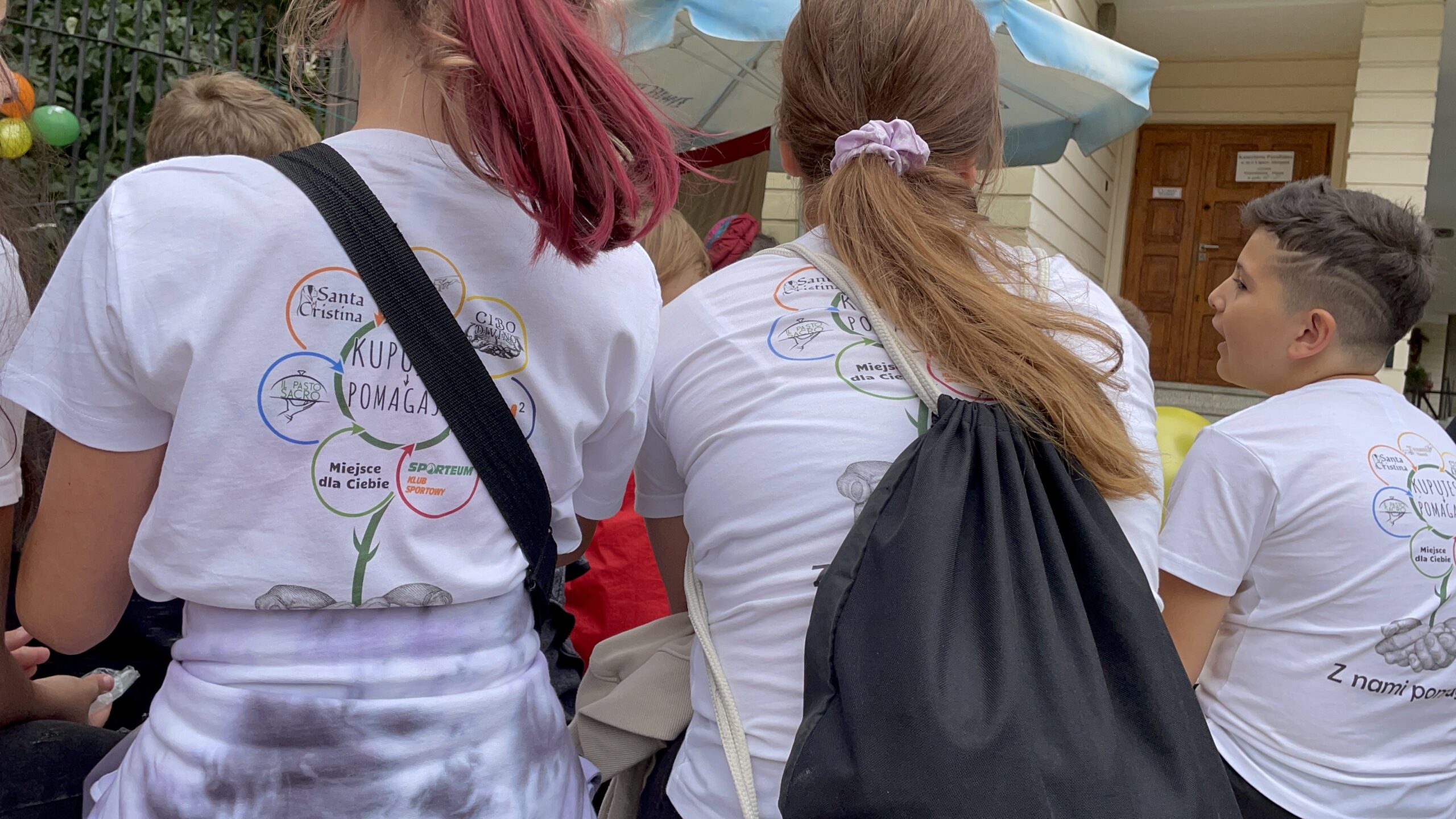Wspólnie w koszulkach akcji. Dzieci i młodzież w białych koszulkach „Kupujesz i pomagasz” przed budynkiem, wśród kolorowych balonów.